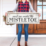 a woman holding a sign that says meet me under the mistletoe