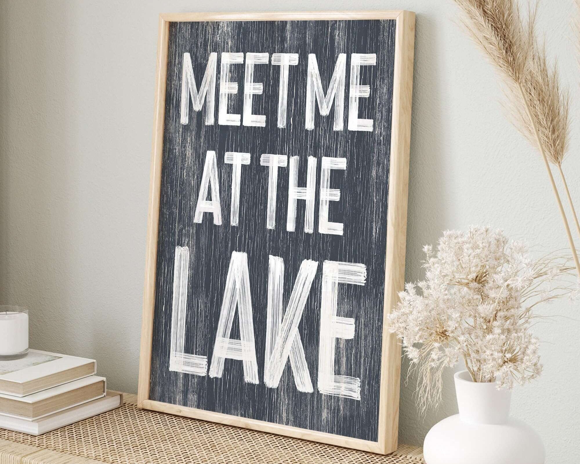 The Meet Me at the Lake Sign in Hale Navy rests on a woven table beside dried flowers and books, making it the ideal lake house decor accent.