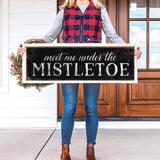 a woman holding a sign that says meet me under the mistletoe