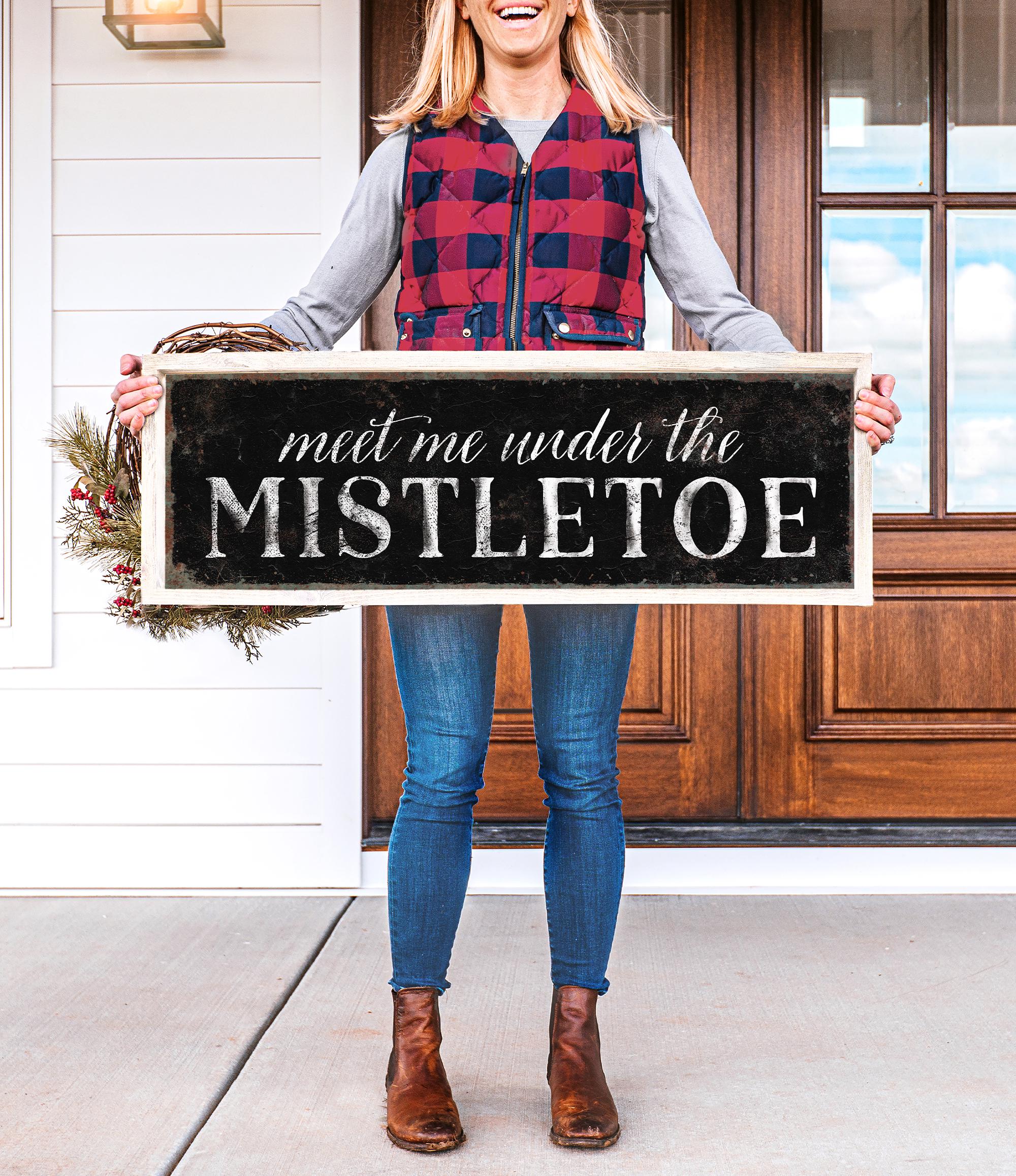 a woman holding a sign that says meet me under the mistletoe