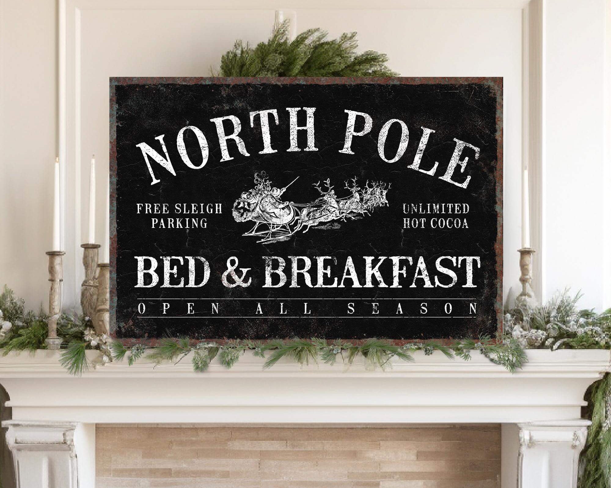 The North Pole Bed & Breakfast Sign, Black, adds festive charm to a mantel with greenery and candles—ideal for modern farmhouse or Christmas wall decor.
